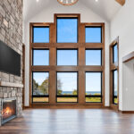 10510 Sugar Creek Great Room + Fireplace. Black interior windows, 2-story great room window wall, vinyl plank floors, ceiling beams, stone fireplace surround with lightweight concrete mantel.