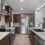 4673 Mill Rd Main Kitchen. Formica "Sea Pearl" Laminate Countertops. Grand Canyon "Sagebrush" Tiled Backsplash. Lone Star Spirit "Ballroom Rodeo" 9" Wide Vinyl Plank Flooring. Cathedral Ceiling. Stained Maple Cabinetry + Woodwork.